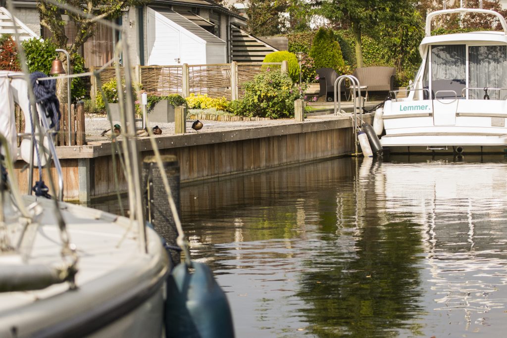 Houten kade met beschoeiing langs waterweg met aangemeerde boten en groene beplanting.