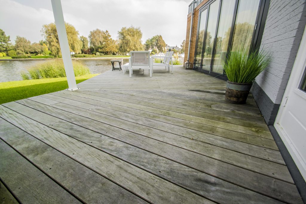 Grijze composiet vlonder aangelegd langs het water met tuinmeubelen en glazen gevel op de achtergrond.