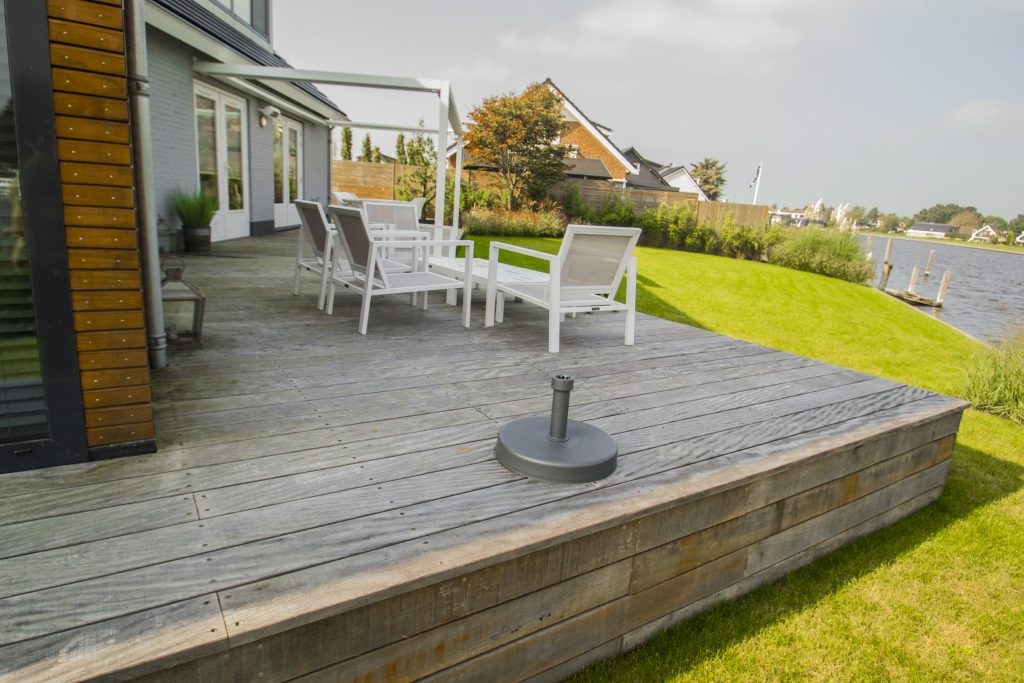 Groot houten terras met composiet vlonders bij moderne woning, met witte tuinmeubelen en groene tuin.