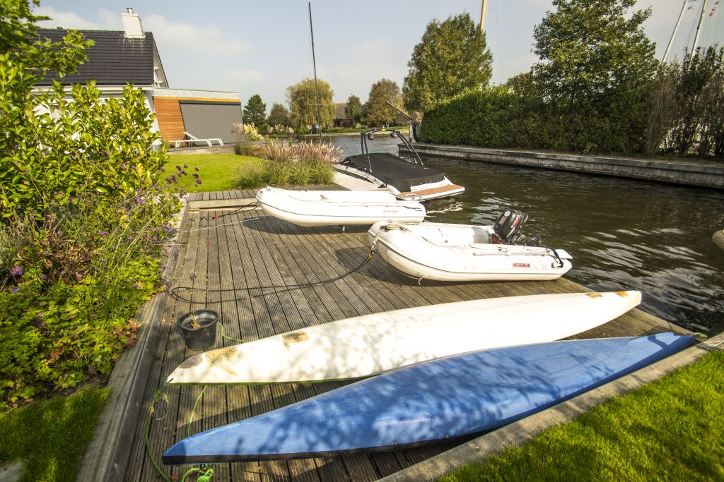 Kleine zeilboten en surfplanken op een houten steiger langs een watergang met groen eromheen.