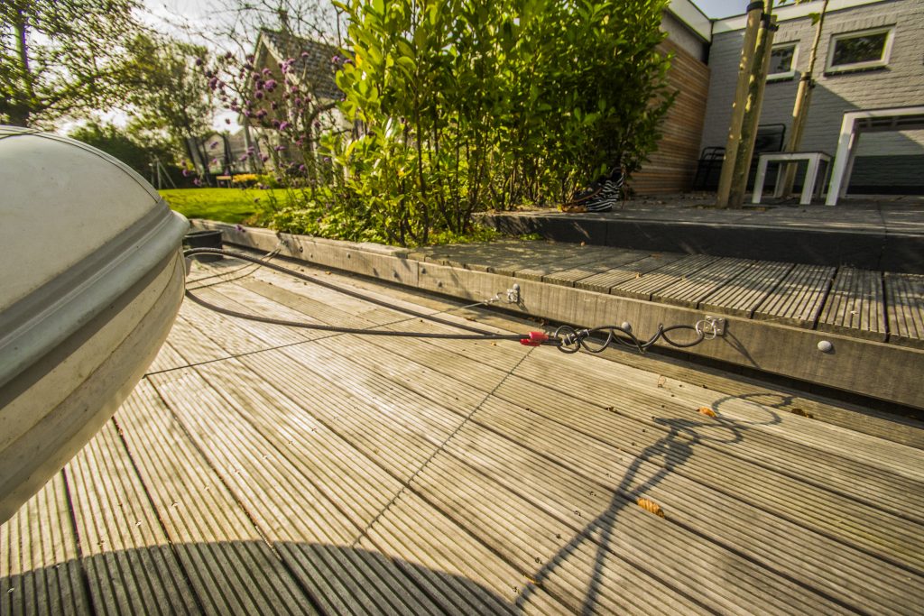 Houten vlonder met kabels en slangen op een terras naast groene begroeiing en houten gevelbekleding.