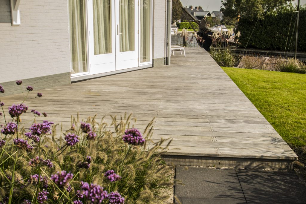 Strak houten terras aangelegd langs witte villa met groen gazon en paarse bloemen op de voorgrond.