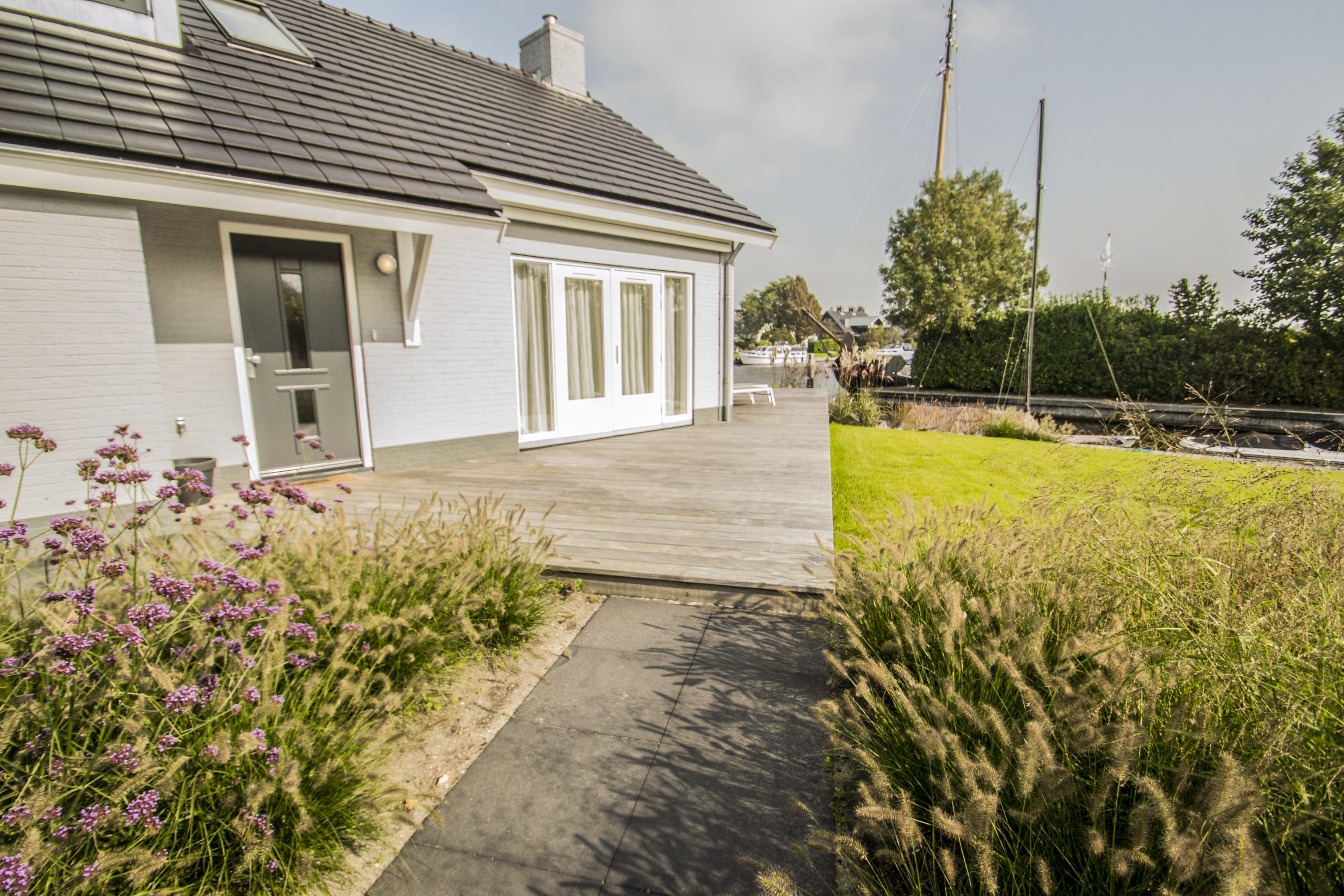 Moderne witte waterwoning met houten vlonderdek, siertuin en groene beplanting langs het water.