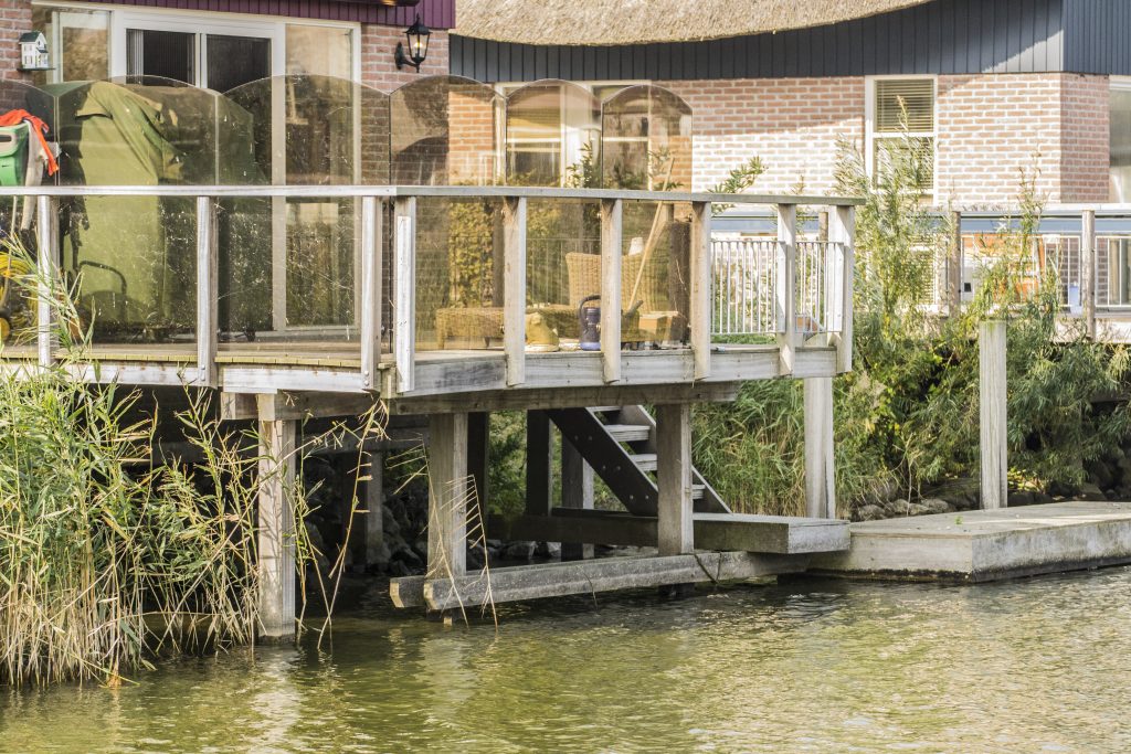 Houten steiger met vlonder aan waterkant, gebouwd op palen naast een woonhuis met glazen veranda.