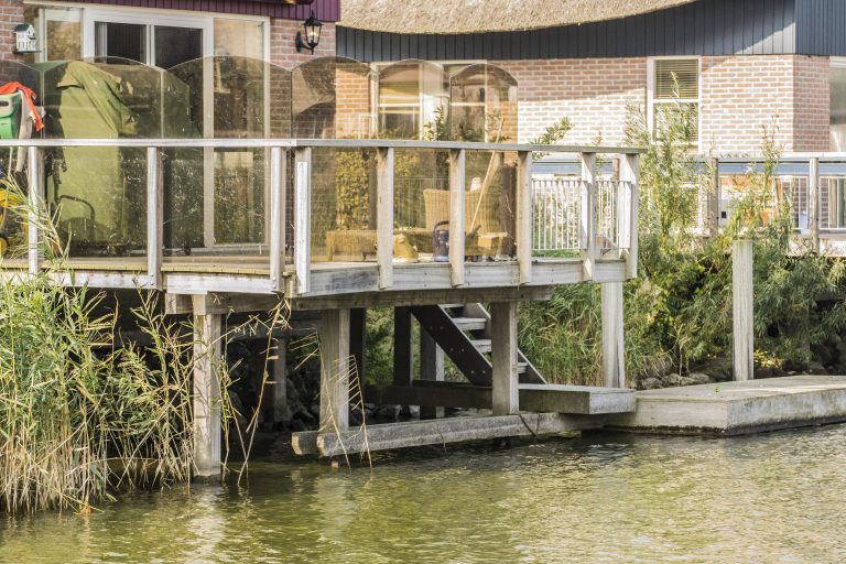 Houten steiger met vlonder aan waterkant, gebouwd op palen naast een woonhuis met glazen veranda.