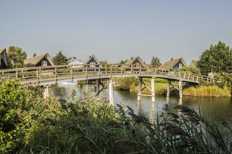 Houten voetbrug over rustig water met rietkraag en vakantiewoningen op de achtergrond.
