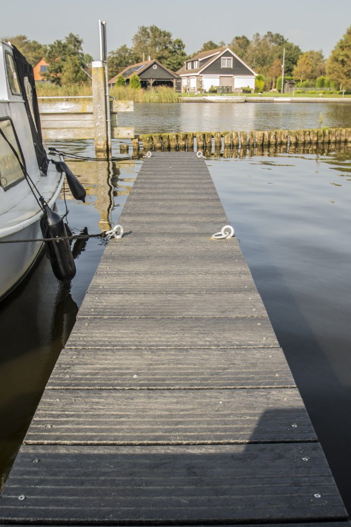 Grijze houten steiger over het water met afgemeerde boot, met op de achtergrond een woonhuis aan het water.
