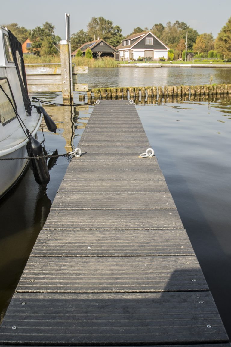 Grijze houten steiger over het water met afgemeerde boot, met op de achtergrond een woonhuis aan het water.