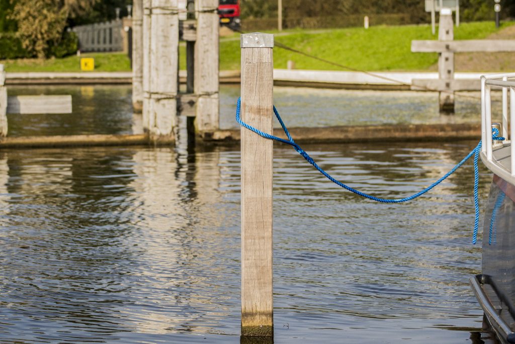 Houten meerpaal met blauwe tros in rustige jachthaven met helder water en sluiswerk op achtergrond.