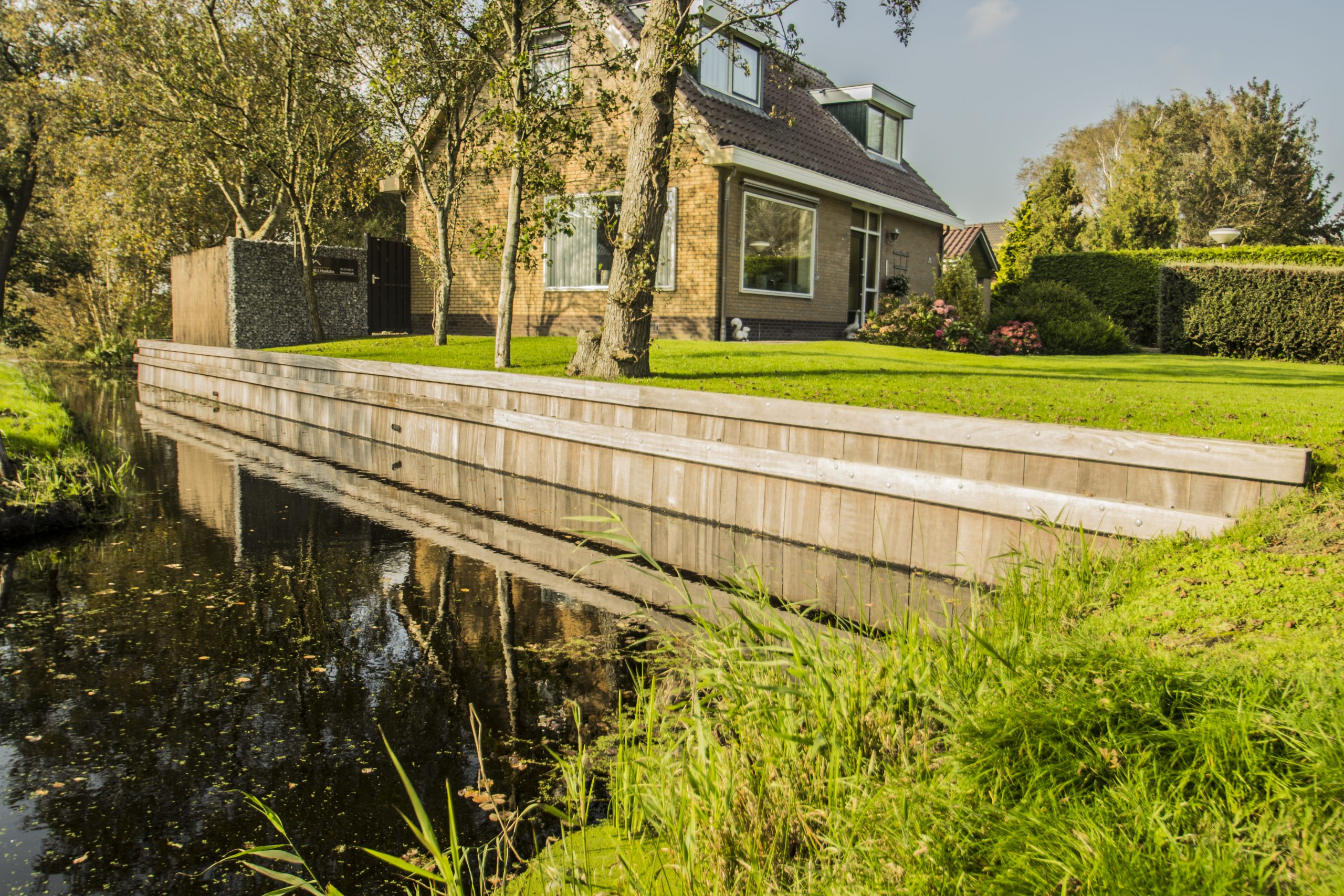 Houten vlonderbrug over sloot bij woonhuis, aangelegd door Bouma Waterwerken in groene tuin.