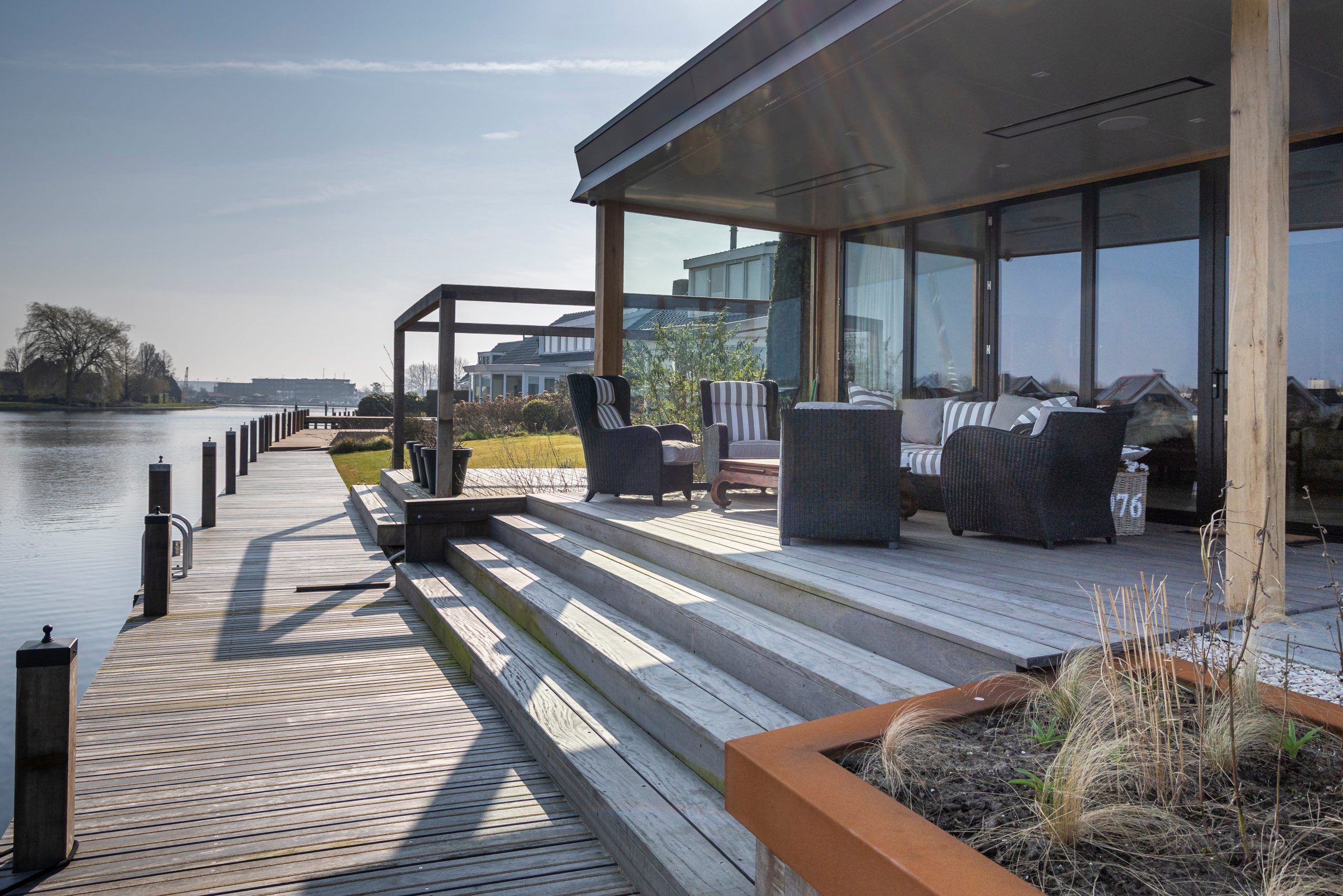 Moderne houten vlonder met loopbrug langs een waterwoning met overdekt terras en loungemeubilair aan het water.