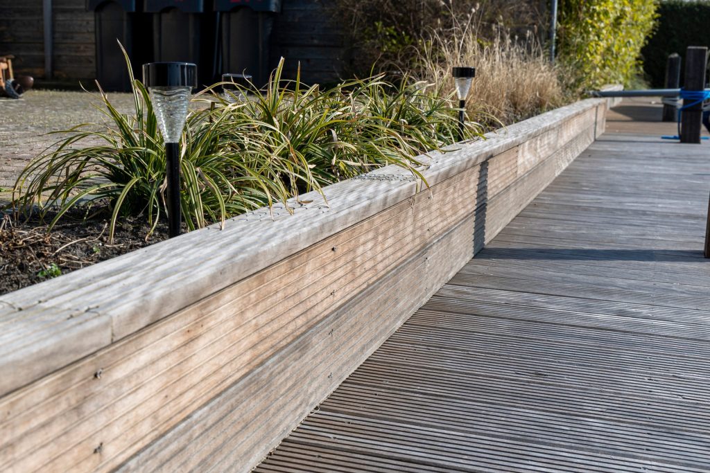 Houten beschoeiing met hardhouten planken langs een vlonder, omgeven door siergras en beplanting.