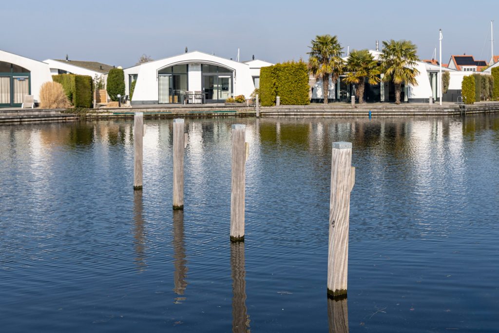 Houten palen in het water langs moderne watervilla's met grote ramen en palmbomen.