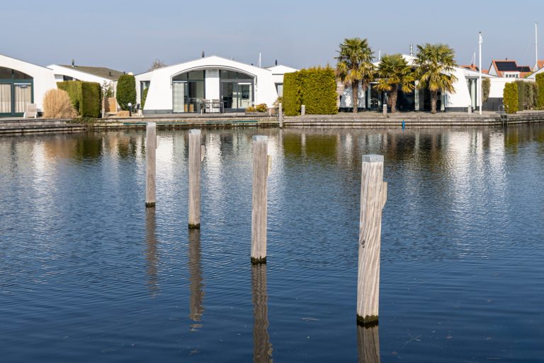 Houten palen in het water langs moderne watervilla's met grote ramen en palmbomen.