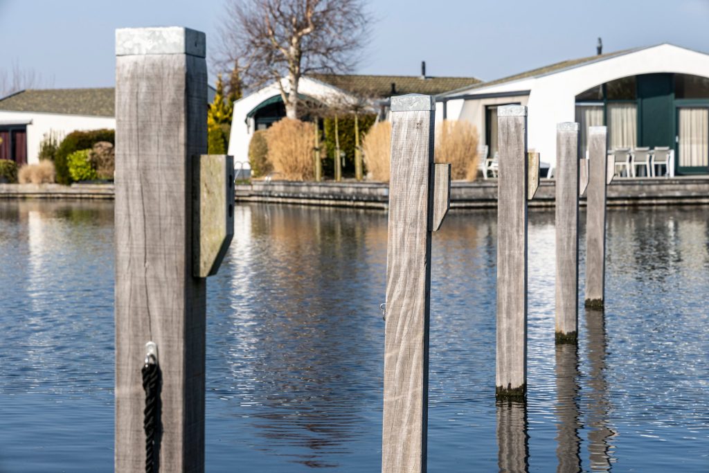 Houten meerpalen in rustig water bij woonhuizen, uitgevoerd door Bouma Waterwerken.