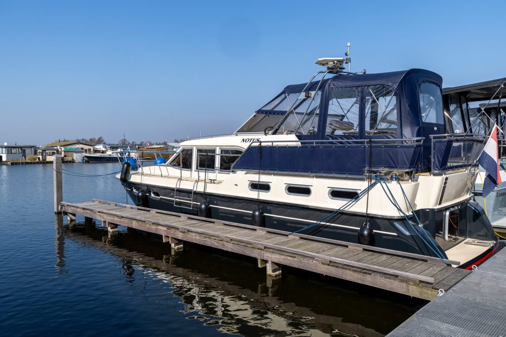 Wit motorjacht met blauwe cabine afgemeerd aan houten steiger op rustig water bij jachthaven.