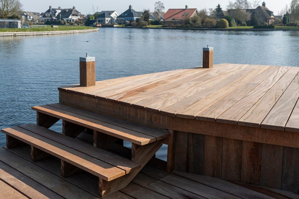 Nieuw aangelegde houten steiger met trap aan het water, omgeven door groen en woningen op de achtergrond.