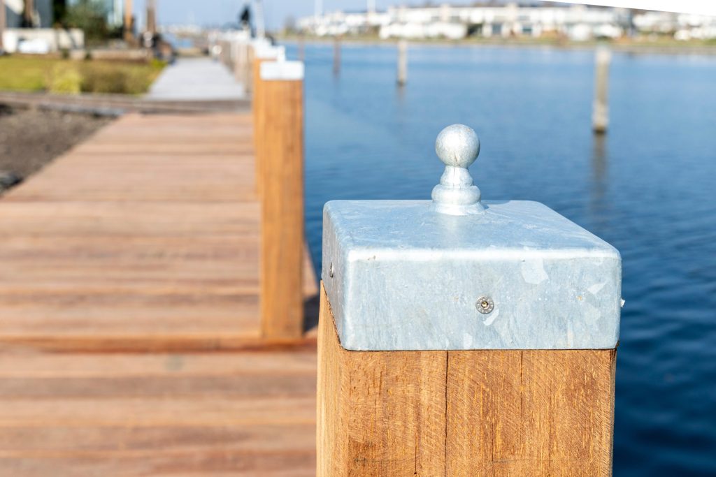 Close-up van een houten steigerpal met gegalvaniseerde metalen afdekplaat en bolkop bij een jachthaven.