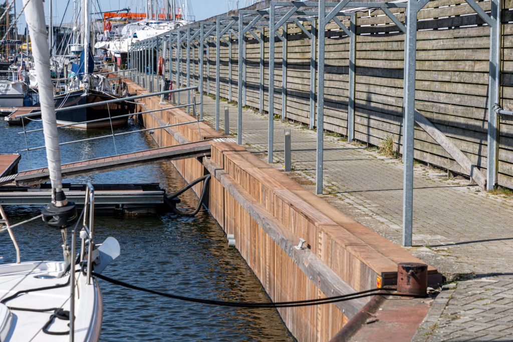 Stalen damwand constructie langs jachthaven met afgemeerde boten en stenen kademuur op de achtergrond.