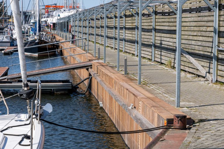 Stalen damwand constructie langs jachthaven met afgemeerde boten en stenen kademuur op de achtergrond.