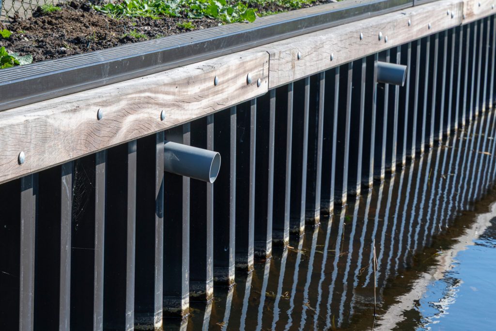 Stalen damwand met houten deksloof langs watergang, inclusief drainage buizen voor waterbeheer.