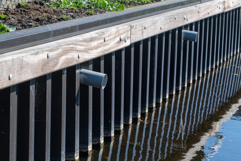 Stalen damwand met houten deksloof langs watergang, inclusief drainage buizen voor waterbeheer.
