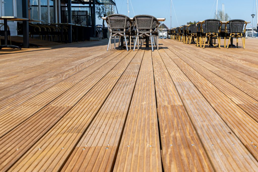 Professioneel aangelegd hardhouten vlonderdek op steiger of terras, met teak planken en buitenstoelen op de achtergrond.