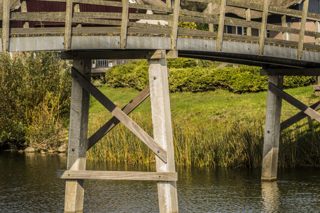 Houten brug met kruisverbindingen op palen boven een waterpartij, omgeven door riet en groen.