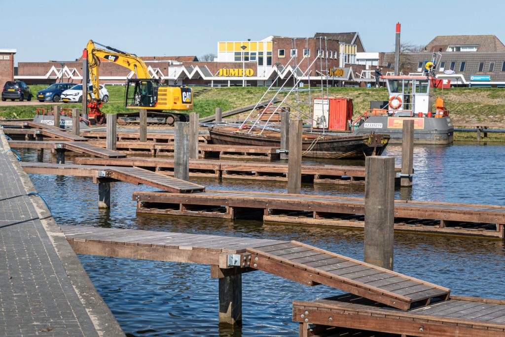 Renovatie van houten steigers in een jachthaven met een graafmachine en werkschip op de achtergrond.